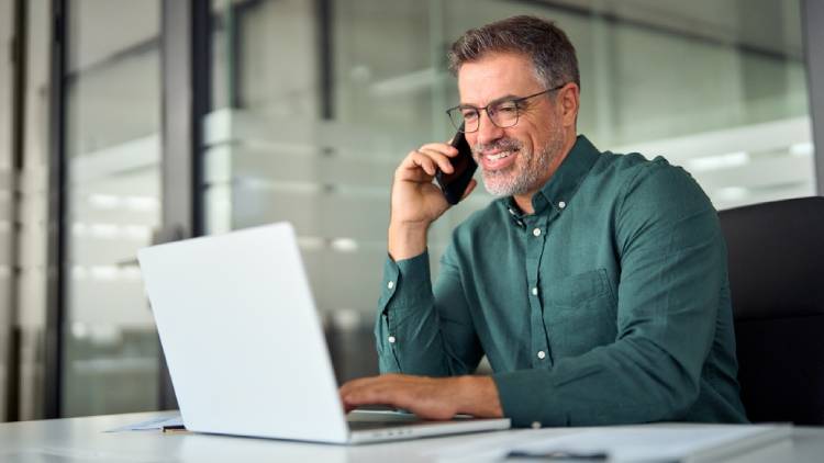 Busy smiling older middle aged business man professional expert or entrepreneur making phone call speaking with client communicating on cellphone using laptop computer sitting at desk in office.