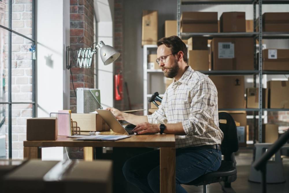 Manager Preparing a Small Cardboard Parcel for Postage. Stylish Young Male Small Business Owner Working on Laptop Computer in Warehouse Facility
